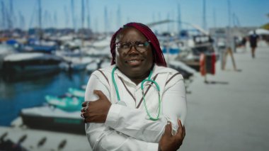 Kolları çapraz duran bir kadın soğuk bir günde arka planda teknelerle marinada titreyen deniz kenarında beyaz önlük ve steteskop giyen bir kadın.