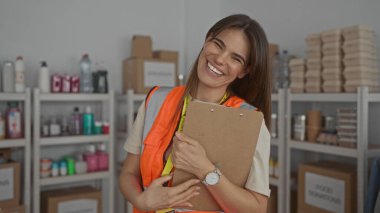 Yansıtıcı yelek giyen bir kadın bağış merkezinde pano tutuyor. Arkaplanda raflar var. Kapalı alanda gönüllü bir çalışma alanı var..