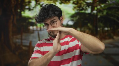 Sakallı, gözlüklü, çizgili tişörtlü, dışarıda yeşil bir parkta mola veren İspanyol bir genç..