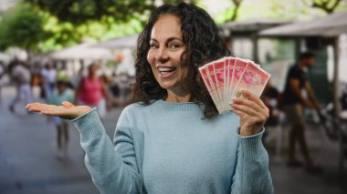 Restoran terasında gülümseyen bir kadın İsrail şekellerini elinde tutarken, bir yandan da paradan söz ediyor ve canlı bir ortamda paranın varlığını vurguluyor..