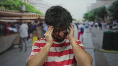 Sakallı, gözlüklü ve kırmızı çizgili tişörtlü bir İspanyol genç kalabalık bir sokakta, arka planda bulanık bir şehir gerginliği hissi uyandırarak strese giriyor..