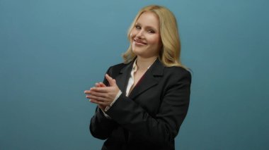 İş elbisesi içinde mavi bir arkaplan karşısında mutlu bir şekilde alkışlayan bir kadın profesyonel bir ortamda pozitiflik ve ifade sergiliyor..
