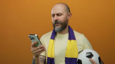 Sakallı bir adam akıllı telefon ve futbol topunu turuncu arka planda tutuyor. Eşarp takıyor. Dalgın görünüyor..