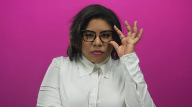 Gözlüklü bir kadın canlı pembe arka planda poz veriyor, beyaz bluz giyiyor, kendine güvenini ve tarzını ifade ediyor, izole edilmiş bir ortamda modern sofistike bir görünüm sergiliyor..