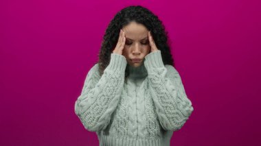 Stresli hisseden kadın, gerilim ve konsantrasyon ifadesiyle çelişen canlı pembe bir arka plana karşı kazak giyerek başını tutuyor..