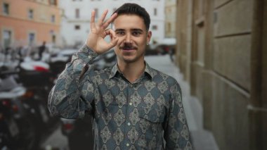 Latin kökenli genç adam şehir manzarası ile çevrili bir şehir caddesinde el hareketi yapıyor..