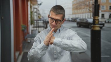 Savunma jesti olarak gözlüklü beyaz bir adam sakin bir sokakta açık şehir ortamında duruyor, hazır olduğunu ve kararlılığını gösteriyor.