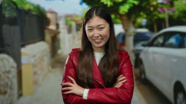 Kırmızı ceketli bir kadın yeşil ve park edilmiş arabalarla çevrili bir caddede gülümsüyor, canlı bir şehir atmosferini canlandırıyor..