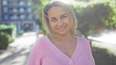 Açık havada gülümseyen kısa sarı saçlı ve pembe kazaklı bir kadın güneşli bir şehir parkı ortamında, yeşilliklerle çevrili, çekici ve neşeli bir hava yayıyor..