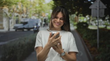 Güneşli şehir caddesinde beyaz gömlek giyerek akıllı telefon kullanan ve arka plandaki bulanık şehir manzarasıyla neşeli iletişimi tasvir eden bir kadın.