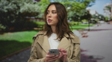 Parkta akıllı telefon kullanan, dikkatle odaklanan, yeşil ağaçlarla çevrili, modern şehir yaşam tarzı ve dijital bağlantı yayan bir kadın.