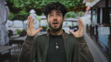 Kafeler ve yeşil ağaçlarla kaplı bir sokakta stresli olduğunu ifade eden sakallı genç bir adam yoğun bir şehir ortamında İspanyol etnik kökeni ve duygusal mücadelesi sergiliyor..