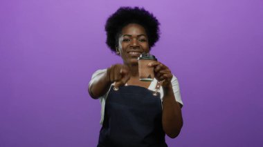 Önlüklü, gülümseyen ve elinde kahve fincanı tutan bir kadın stüdyo ortamında canlı mor bir arkaplan, pozitiflik ve nişan sergiliyor..