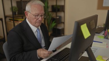 Modern ofis içinde bir bilgisayarda çalışan takım elbiseli kıdemli bir adam, belgeleri okumaya ve görevleri düzenlemeye odaklanmış..