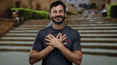 Dışarıda sakallı bir adam gülümsüyor ve bir şehir caddesinde kendini kucaklıyor, huzur ve mutluluk taşıyor..