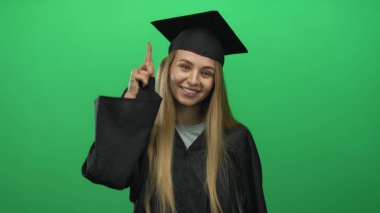 Mezuniyet üniformalı genç beyaz kadın yeşil arka planda kutlama yapıyor, neşeli bir ifadeyle, başarıyı ve başarıyı sembolize ediyor..