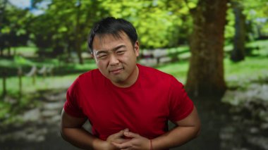 Kırmızı tişörtlü bir adam, yemyeşil yemyeşil bir parkta midesi ağrıyan bir şekilde sıkışıyor. Açık havada rahatsızlığı vurguluyor..