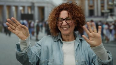 Dışarıdaki Vatikan ortamında gülümseyen bir kadın, arka planda insanlarla birlikte St. Peter Meydanı 'nda hareketli bir şehir atmosferi sergiliyor..