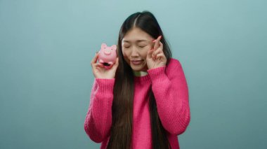 Asyalı bir kadın domuz kumbarasını mavi arka planda umut içinde tutuyor pembe bir kazak giyiyor ve finansal iyimserliğini dile getiriyor..