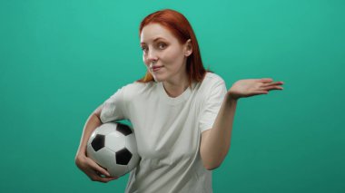 Futbol topunu tuhaf bir ifadeyle tutan kadın yeşil bir zemine karşı duruyor. Günlük beyaz bir gömlek giyiyor. Düşünceli ve enerjik bir görünümü var..