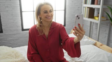 Kırmızı pijamalarıyla gülümseyen bir kadın modern yatak odasında selfie çekiyor. Parlak pencereli, rahat bir yatak odası, rahat bir sabah rutini ve neşeli bir iç mekan..
