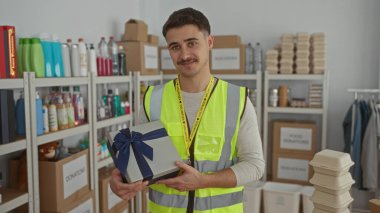 Yansıtıcı yelek giyen genç bir İspanyol, bağış merkezinde bir hediye kutusu tutuyor. Etrafında bir sürü malzeme var..