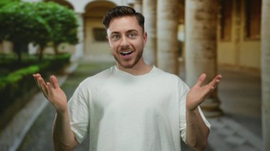 Sakallı bir adam güvenli bir şekilde şehir ortamında gülümsüyor, tarihi mimariyle çevrili, beyaz gömlek giyiyor, rahat ve enerjik tavırlar sergiliyor..