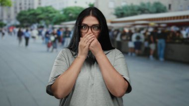 Gözlüklü genç bir kadın marketin dışındaki işlek şehir sokağında dikiliyor. Elleriyle ağzını kapayarak şehrin bulanık arka planına şaşırıyor..
