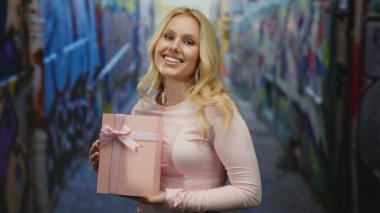 Açık havada elinde pembe hediye kutusuyla gezen bir kadın. Grafiti arka planında pembe kıyafetler giyiyor. Uzun sarı saçlı, hayat dolu şehir atmosferinde gülümsüyor.