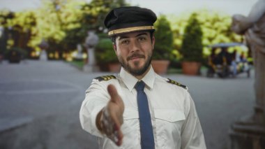 Sakallı ve yüzbaşı üniformalı genç adam gülümsüyor ve açık hava parkında arka planda bulanık ağaçlarla el ele tutuşuyor.