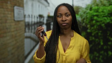 Şehir caddesinde elinde araba anahtarlarıyla duran, parlak sarı gömlekli, kendine güvenini ve bağımsızlığını canlı bir şehir ortamında sergileyen bir kadın..