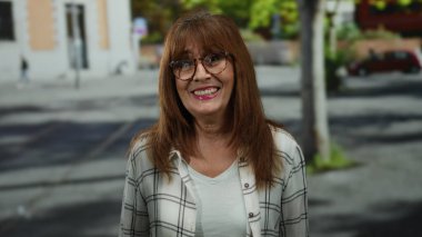 Bir kadın şehir caddesinde, gözlüklü ve ekose gömlekli bir şekilde gülümseyip kendini işaret ederek, İspanyol mahallesinde neşeli bir açık hava sahnesi yaratıyor..