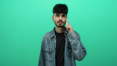 Kot ceketli genç adam akıllı telefondan konuşuyor, parlak yeşil arka planda izole edilmiş..