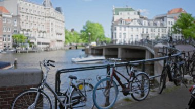 Amsterdam 'da bir kanal boyunca park etmiş bisiklet manzarası bulanık. Arka planda geleneksel Hollanda mimarisi ve köprünün altından geçen bir tekne var..