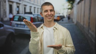 Sokakta el kol hareketi yapan bir adam şehir ortamında kaldırılmış, günlük giysiler giymiş ve arka planda bulanık arabalarla kameraya gülümsüyor..