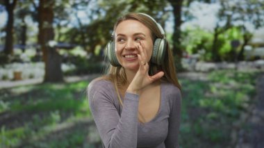 Kulaklıklı bir kadın, yeşil bir parkta konuşuyor ve gülümsüyor. Etrafı yeşilliklerle çevrili. Sırrını paylaşıyor, neşeli ve ilgi çekici bir sahne yaratıyor..