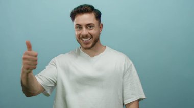 Parmakla gülümseyen genç bir adam canlı mavi bir arka plana karşı bir jest, izole edilmiş bir stüdyo ortamında pozitiflik ve güven yayıyor.