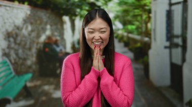 Kentsel sokak ortamında gülümseyen bir kadın pembe giyinmiş, yeşillikle mutluluğu ifade eden ve parlak bir günde bulanık arka planda oturan insanlar..