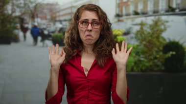 Kıvırcık saçlı ve gözlüklü bir kadın şehir sokaklarında duygularını ifade ediyor, kırmızı bir gömlek giyiyor, arka planda bulanık yayalar olan bir şehir arka planına karşı..