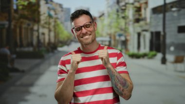 Kenar mahallede kendine güvenen genç bir adam kırmızı ve beyaz çizgili bir gömlek giyerken iki parmağını kaldırıyor..