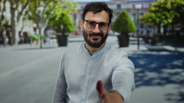 Gözlüklü İspanyol adam, güneşli bir sokakta, arka planda modern bir şehir ortamında yeşil ağaçların oluşturduğu dostça bir selamlaşmayla elini uzatıyor..