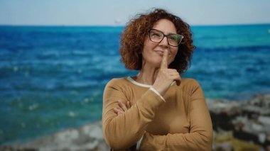 Orta yaşlı İspanyol bir kadın, deniz kenarında, mutlu bir ifadeyle, gözlük takıyor ve kahverengi bir süveter giyiyor, canlı mavi bir deniz ve gökyüzünün zeminine karşı..