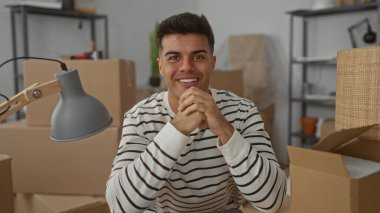 Yeni ev ortamında kutuları taşıyarak gülümseyen genç adam, yeni döşenmiş bir dairede yeni bir başlangıç öneriyor..