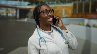 Kentsel bir bölgede, açık havada gülümseyen ve konuşan bir kadın, gözlük ve steteskop takıyor, profesyonellik ve bağlantı duygusunu iletiyor..