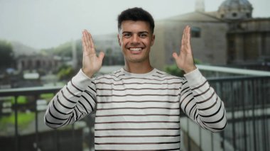 Roma harabelerinin önünde poz veren genç bir adam, gün boyunca açık havada gülümseyen, çizgili bir kazak giyen, kentsel bir ortamda enerjik ve çekici bir enerji sergileyen..