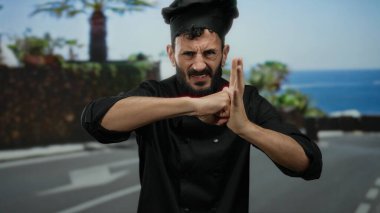 Siyah üniformalı İspanyol şef, güneşli bir sokakta yumruklarıyla enerji dolu jestler yapıyor. Palmiye ağaçları ve mavi gökyüzü arkaplanı arasında kararlılık ve duyguyu gösteriyor..