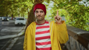 Kırmızı bereli ve sarı ceketli genç adam şehir sokağında duruyor, baş parmağıyla aşağı işaret ediyor, arka planda canlı yeşil ağaçlar ve park edilmiş arabalarla çevrili..