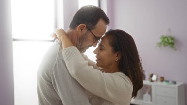 İspanyol çift, sakin bir oturma odasında sevgi dolu bir şekilde kucaklaşırken orta yaşlı aile hayatının sıcaklığını ev içinde erkek ve kadınla paylaşıyorlar..