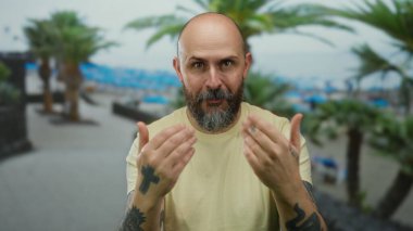 Kel sakallı adam sahilde el kol hareketleri yapıyor. Arka planda palmiye ağaçları ve deniz var. Dostane ve davetkar bir atmosferi var..