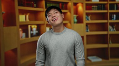 Kütüphanede arka planda rafları olan genç bir adam, kapalı bir oda ortamında neşeli bir ifade sergiliyor, çağdaş bir gündelik tarzı sergiliyor..